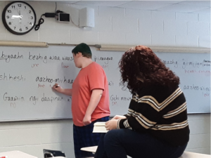 A student in a red shirt and blue jeans writes in a native language on a white board in a classroom. A teacher looks on wearing a striped sweater and black pants. The teacher is sitting on a desk.