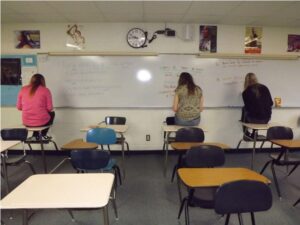 Three students sit on top of desks in front of a white board in a classroom.